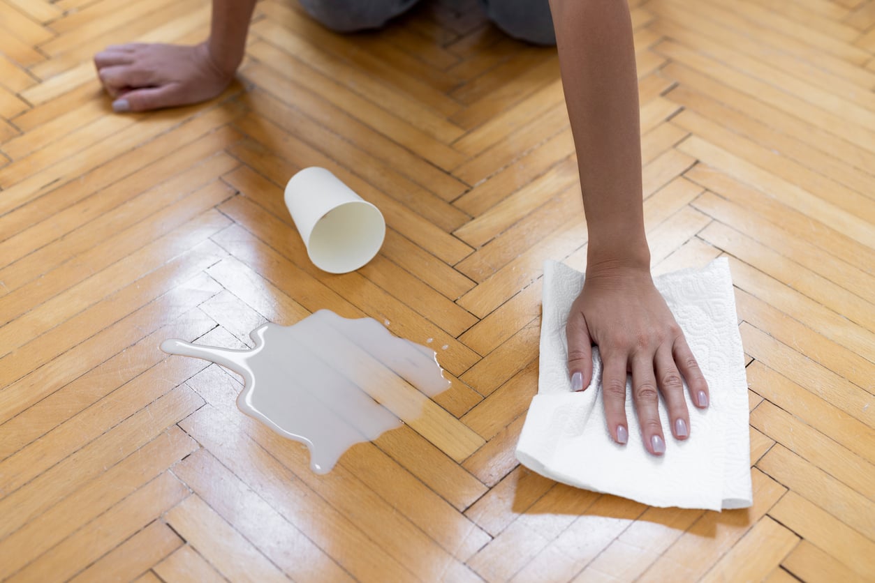 A person wipes up a puddle of spilled liquid on a wooden floor with a paper towel next to an overturned paper cup.