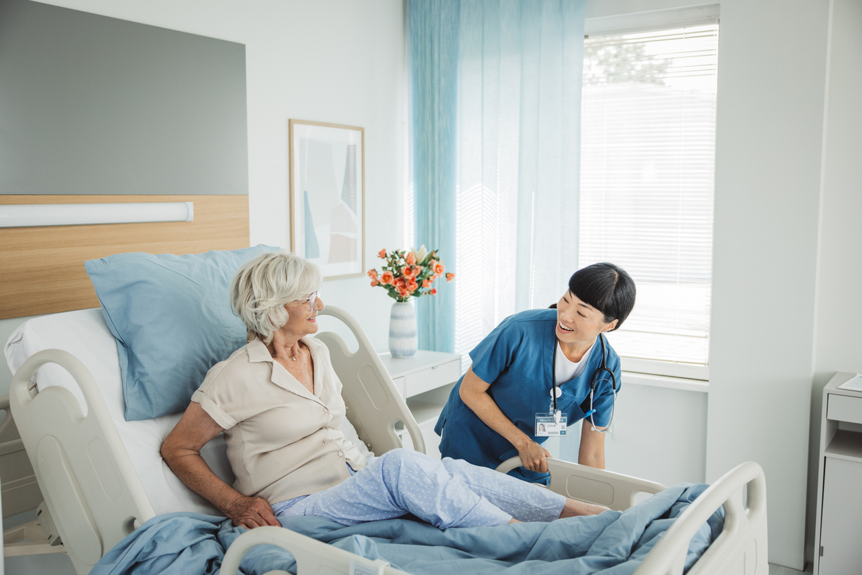 A nurse in blue scrubs smiles while talking to an elderly patient sitting on a hospital bed.
