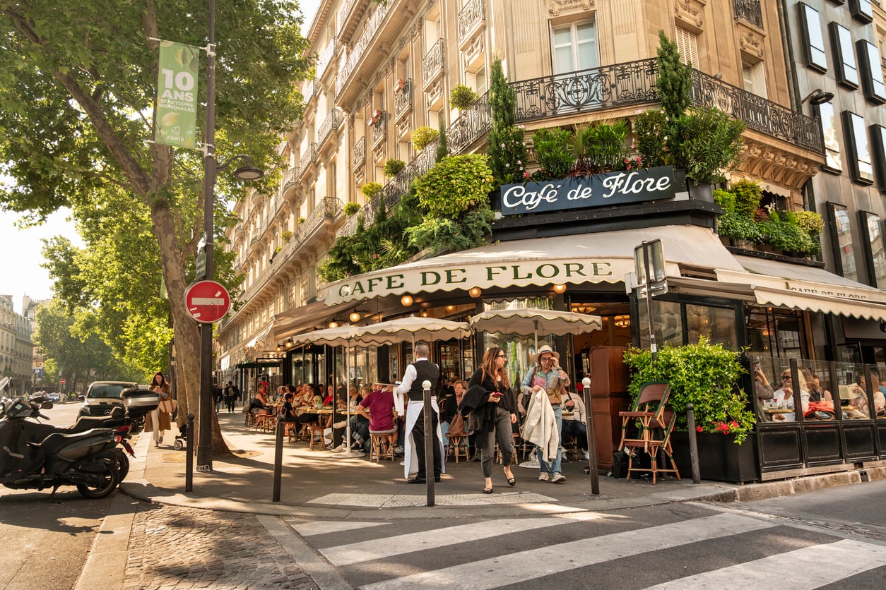 People seated at tables on the sidewalk patio of the Café de Flore in Paris, with a server standing at the entrance.