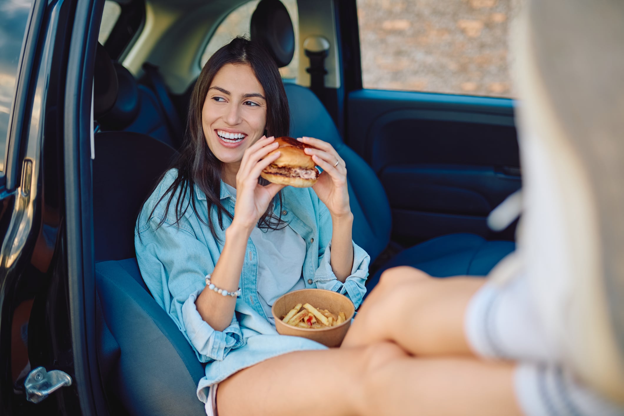 A smiling woman sits in the passenger seat of a car, holding a burger with a container of french fries on her lap.