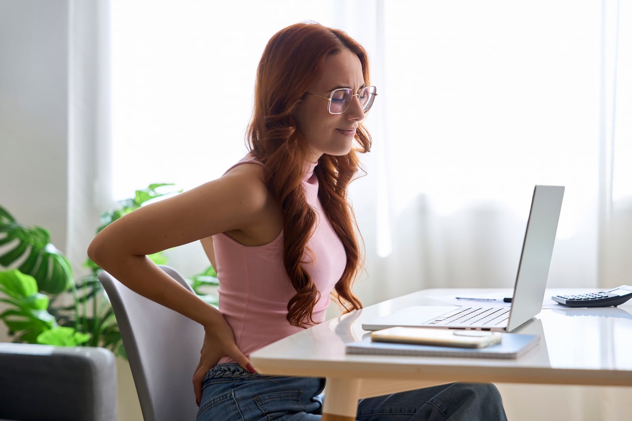 A woman sitting at a desk with a laptop winces while holding her lower back.