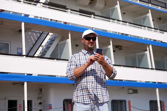A man wearing a white baseball cap, sunglasses, and a plaid shirt holds a smartphone while standing on the deck of a cruise ship.