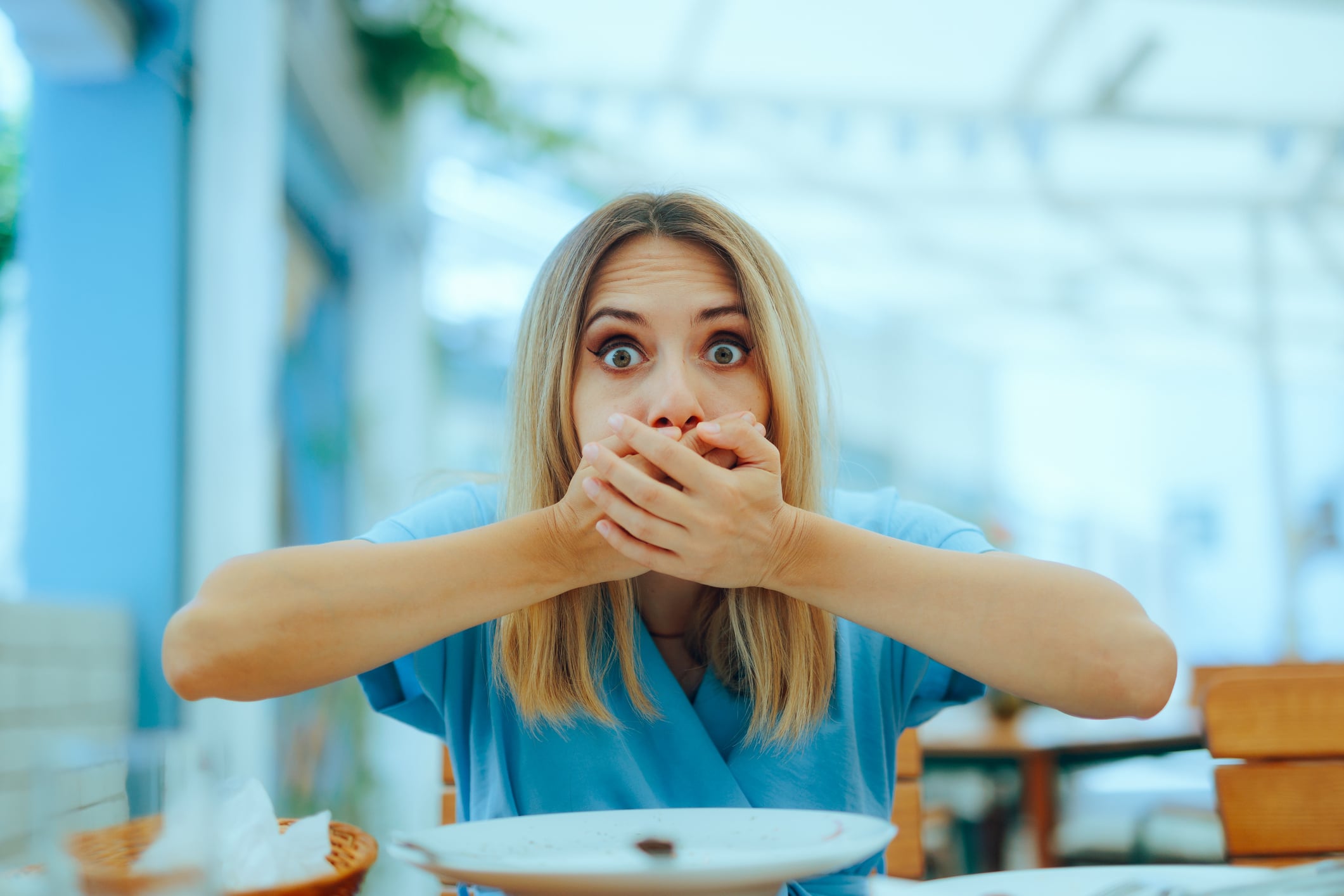 A woman sitting at a table with a wide-eyed, shocked expression while covering her mouth with both hands.