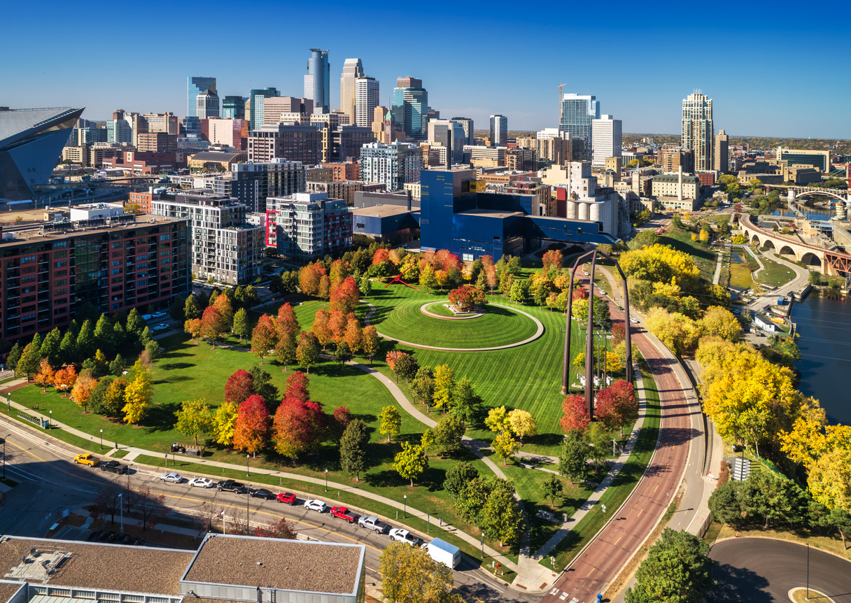 Aerial view of a park with fall foliage, a circular lawn, and steel sculptures, set against the Minneapolis city skyline.