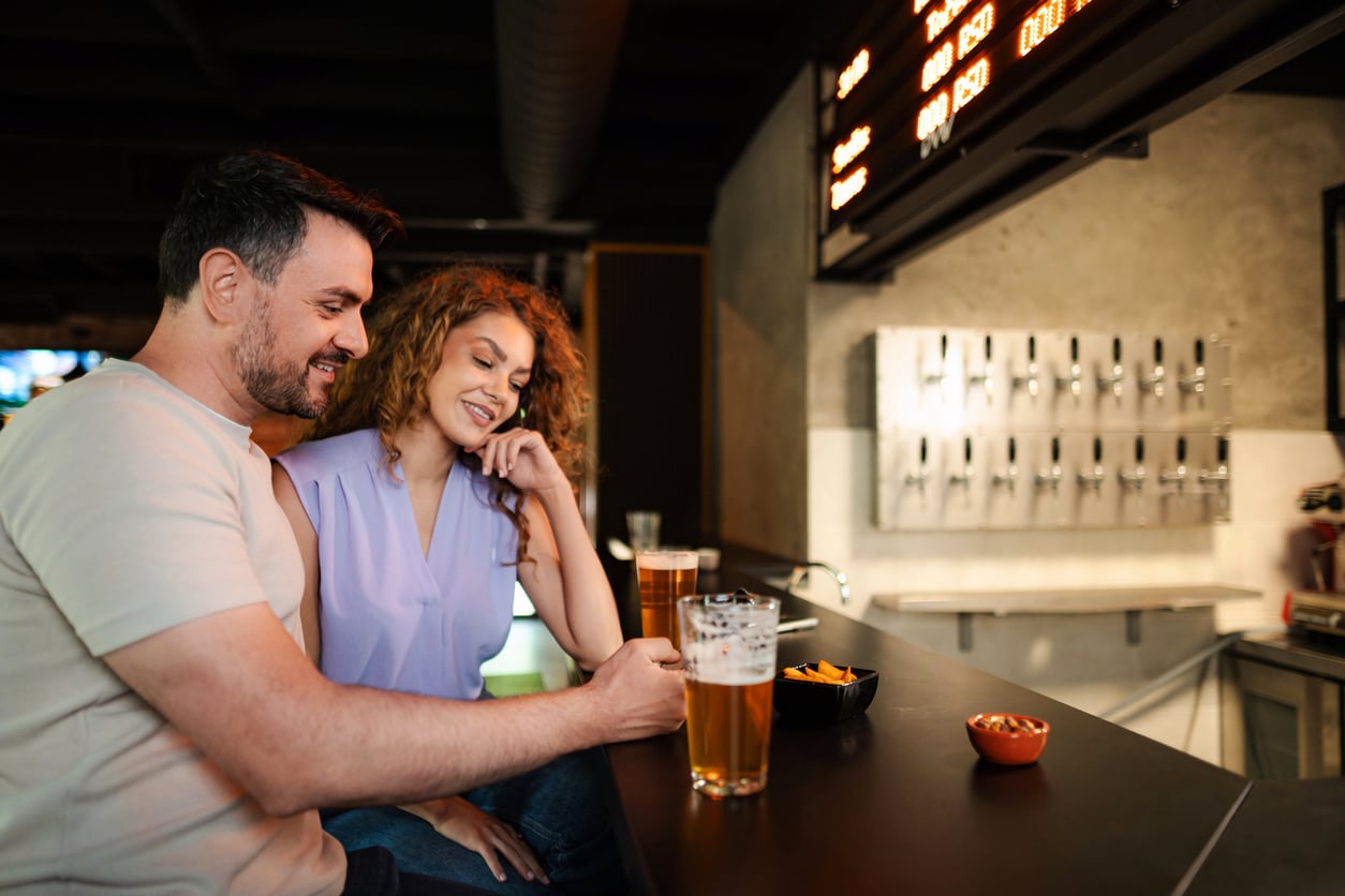 A man and woman sit at bar seating with glasses of beer and small bowls of snacks.