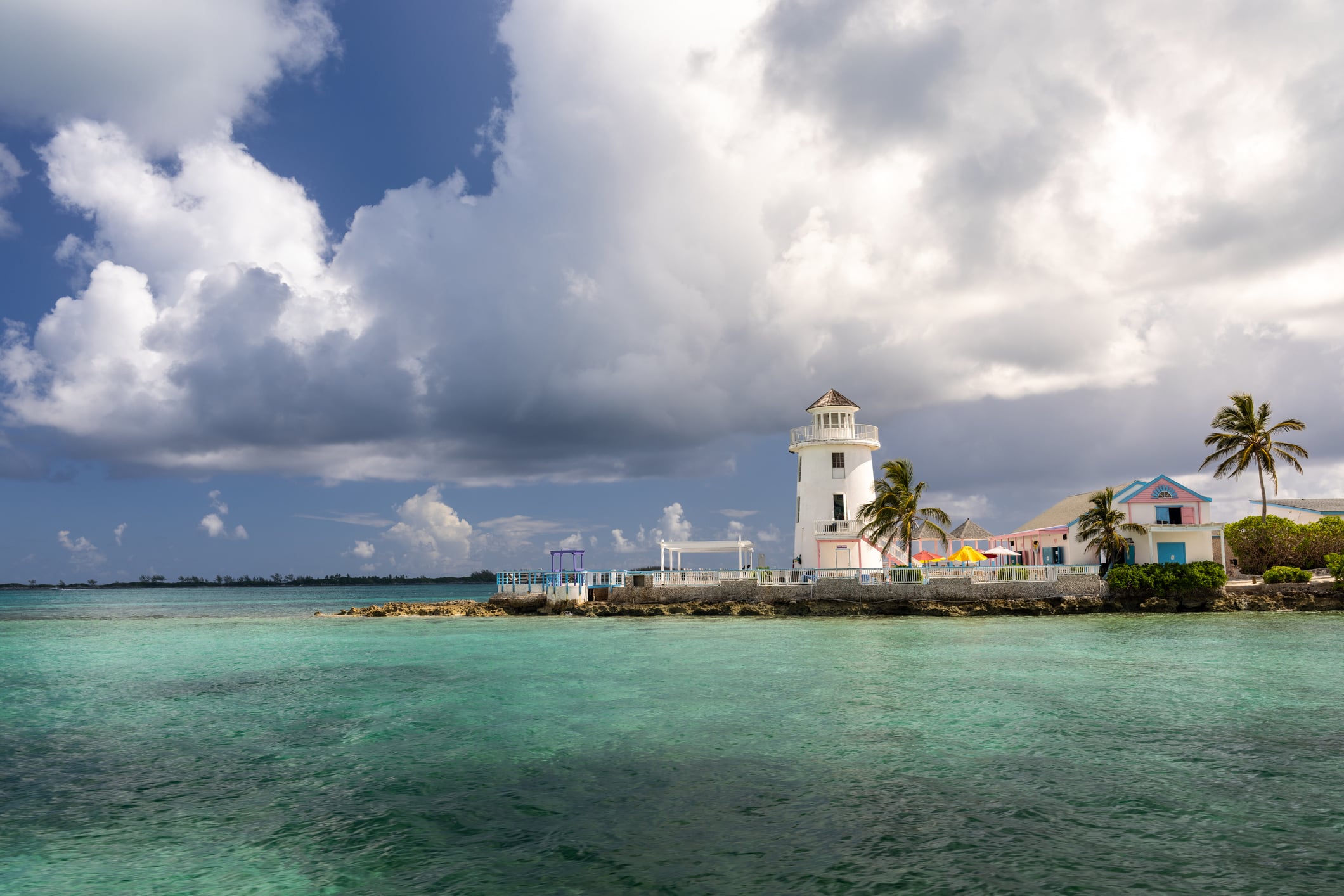 A white lighthouse and adjacent pastel buildings on a rocky point surrounded by turquoise water under a partly cloudy sky.
