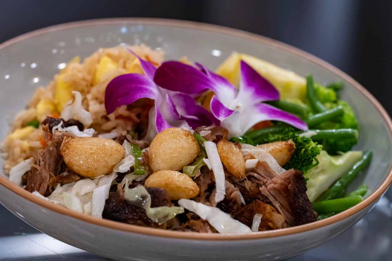 A bowl containing crispy pork, fried rice, green beans, broccoli, and whole garlic cloves, garnished with a purple orchid.