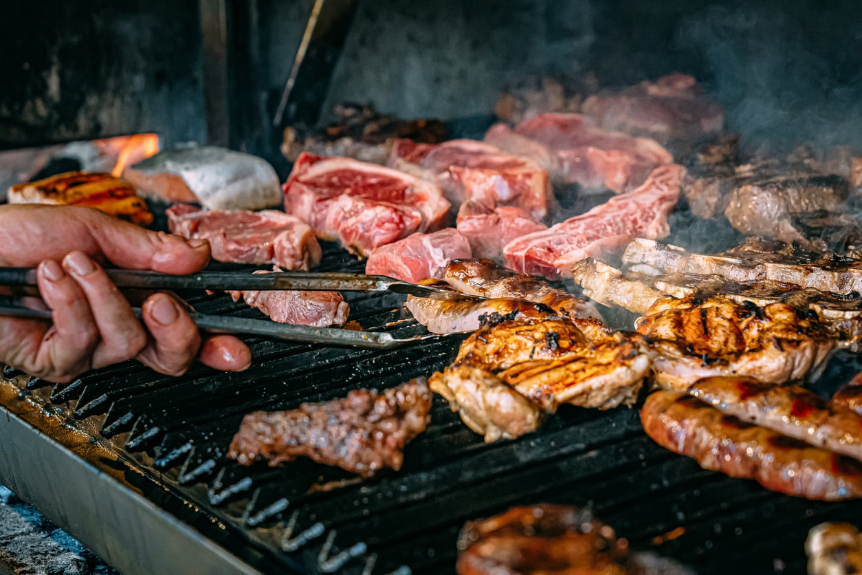 A hand uses metal tongs to flip pieces of raw and grilled meat on a charcoal grill.
