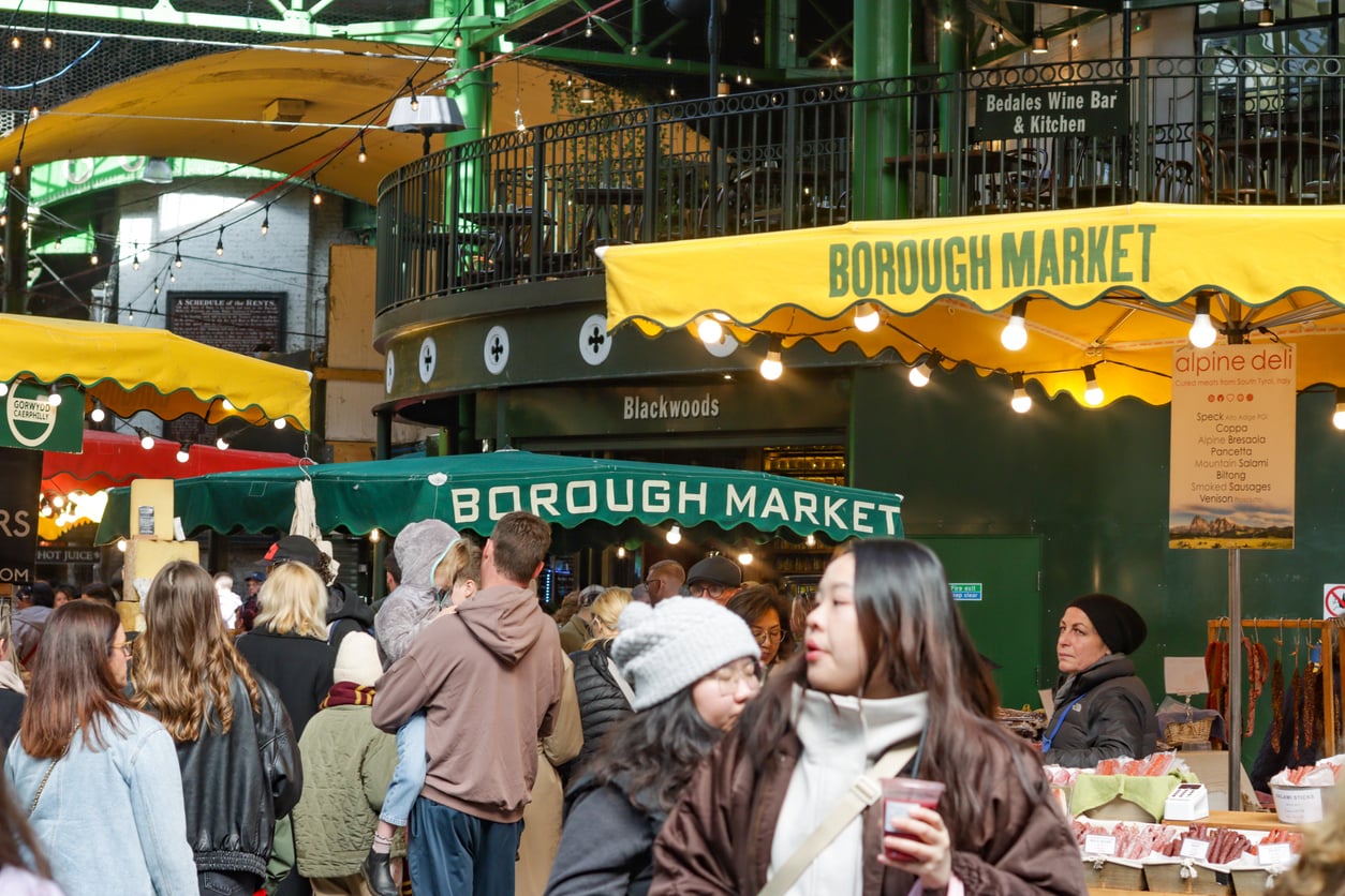 A crowded Borough Market scene with patrons browsing stalls, including an Alpine Deli counter featuring a list of cured meats.