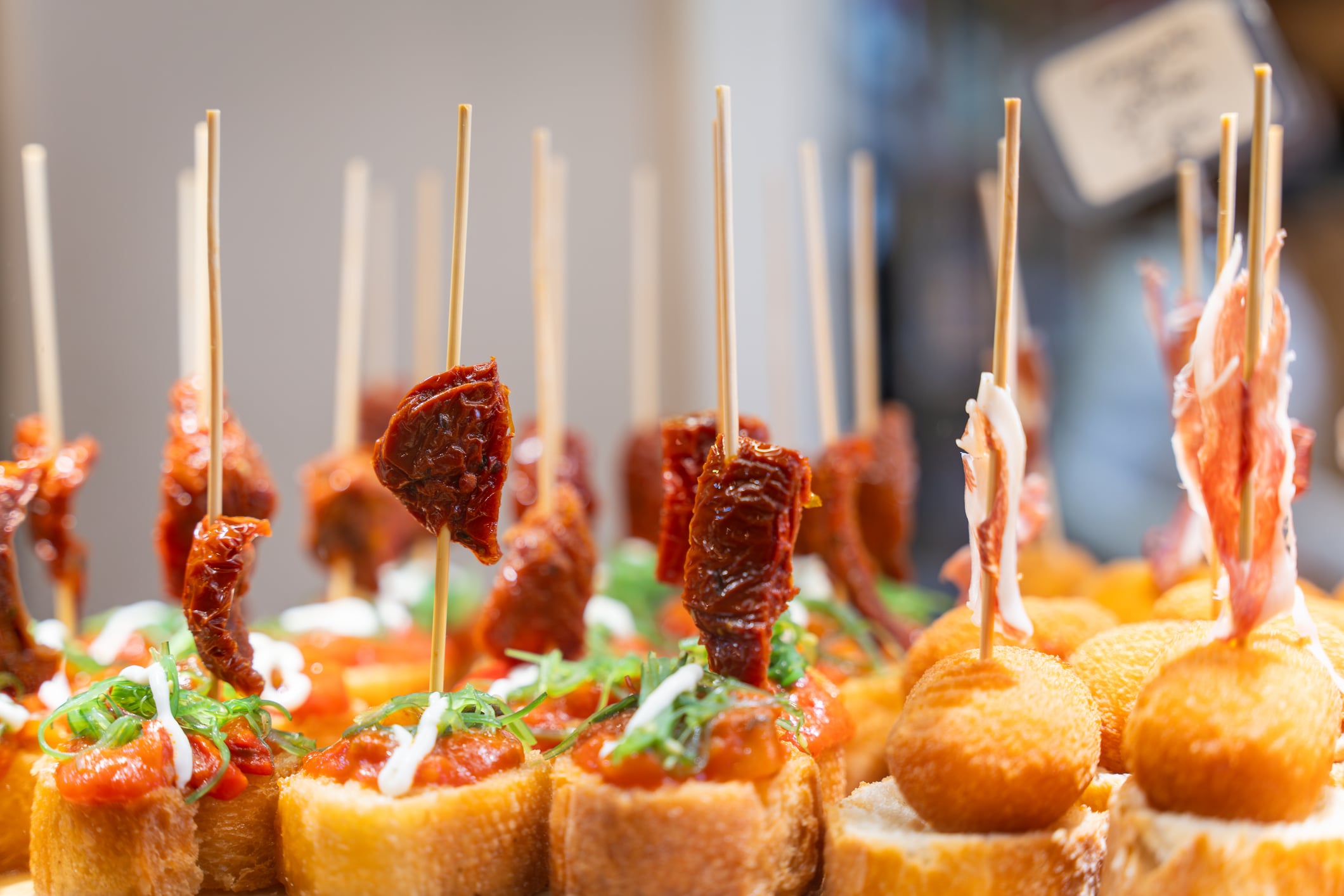 Assorted tapas served on bread slices with skewers, topped with sun-dried tomatoes, herbs, or thin slices of cured meat.