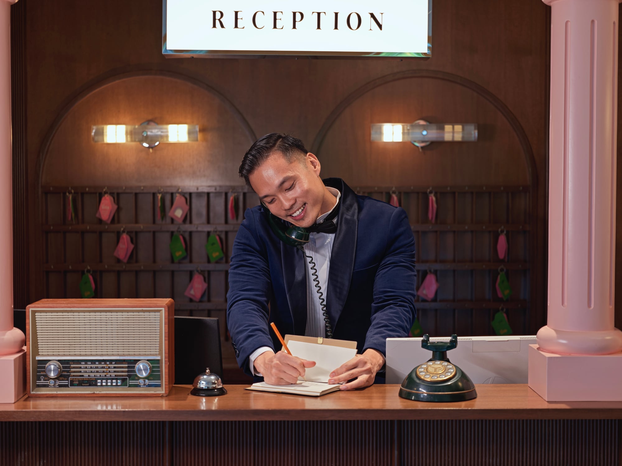 A receptionist in a blue blazer speaks on a vintage telephone while writing in a notebook behind a hotel reception desk.