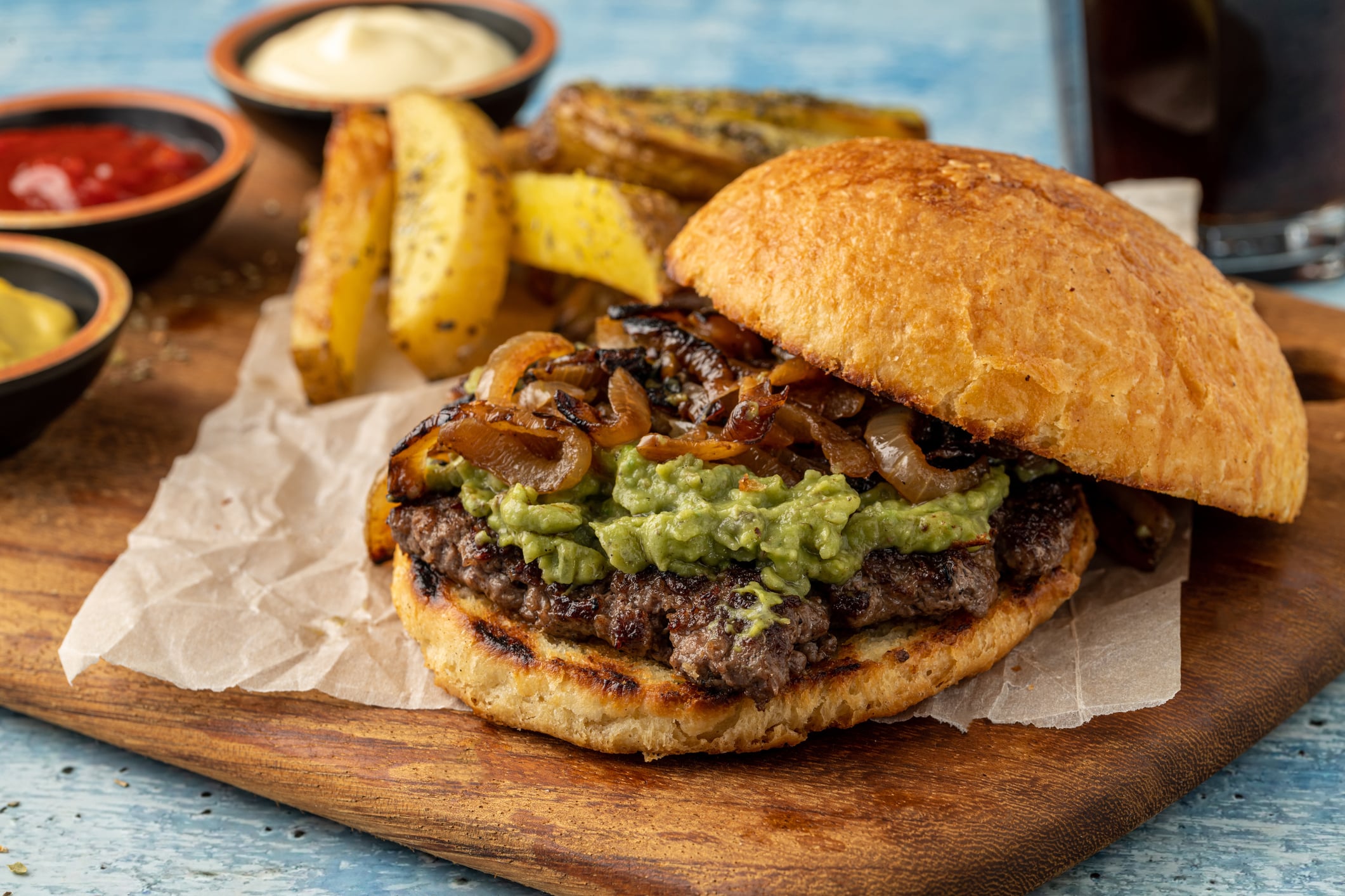 A burger topped with guacamole and caramelized onions on a wooden board, served with potato wedges and dipping sauces.