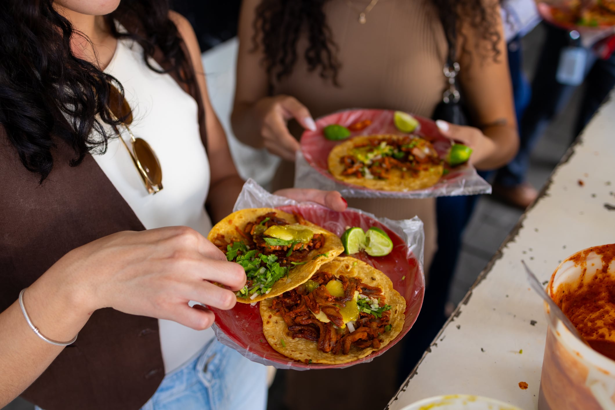 Two people holding plastic plates filled with tacos topped with cilantro and lime wedges.