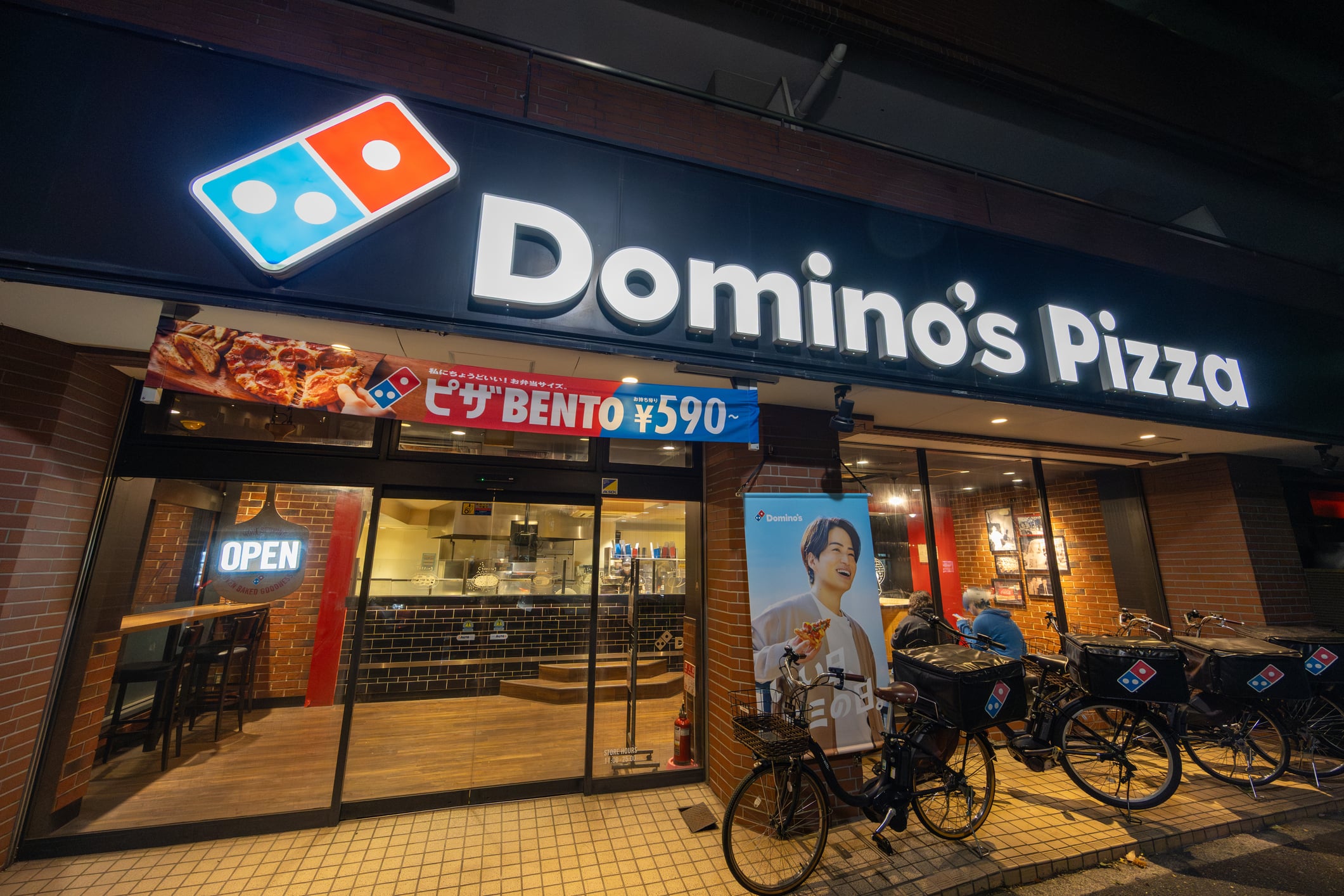 Exterior view of a Domino’s Pizza storefront at night, with an “Open” sign in the window, a banner advertising a 590 yen bento meal, and delivery bicycles parked out front.