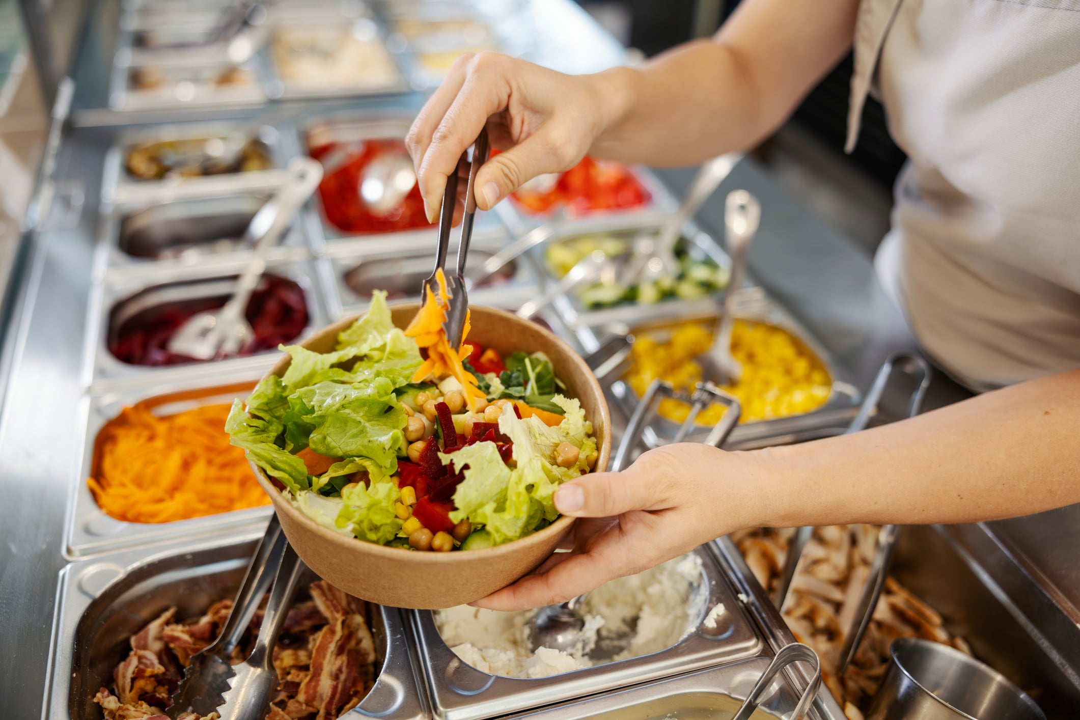 A person using tongs to add shredded carrots to a salad bowl at a food prep station.