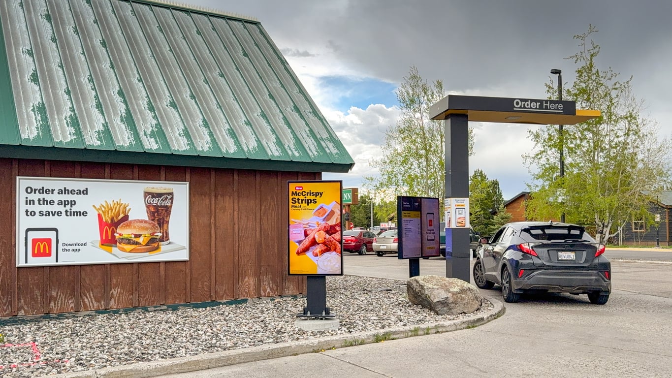 A car parked at a McDonald’s drive-thru, with menu signs featuring a burger meal and crispy chicken strips displayed on the building and stand.