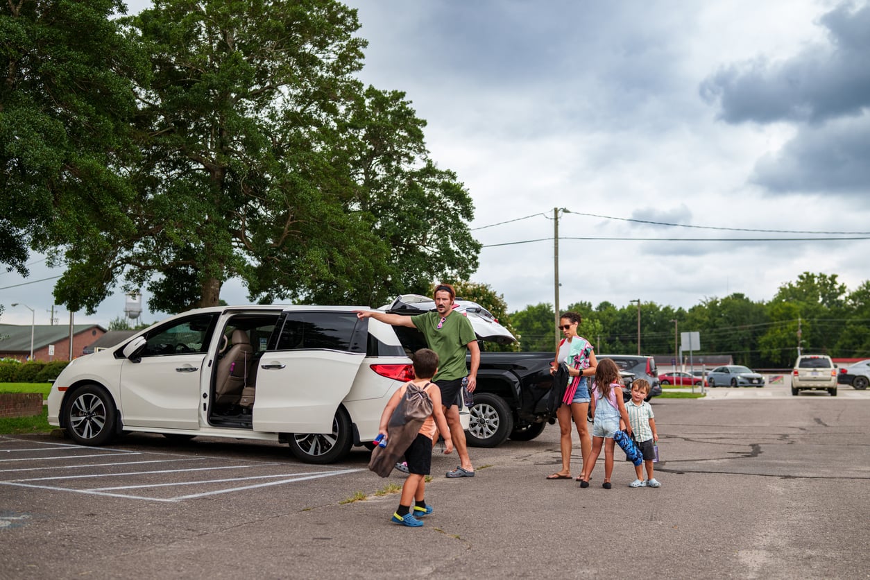A man and a woman stand by an open minivan in a parking lot with three children.