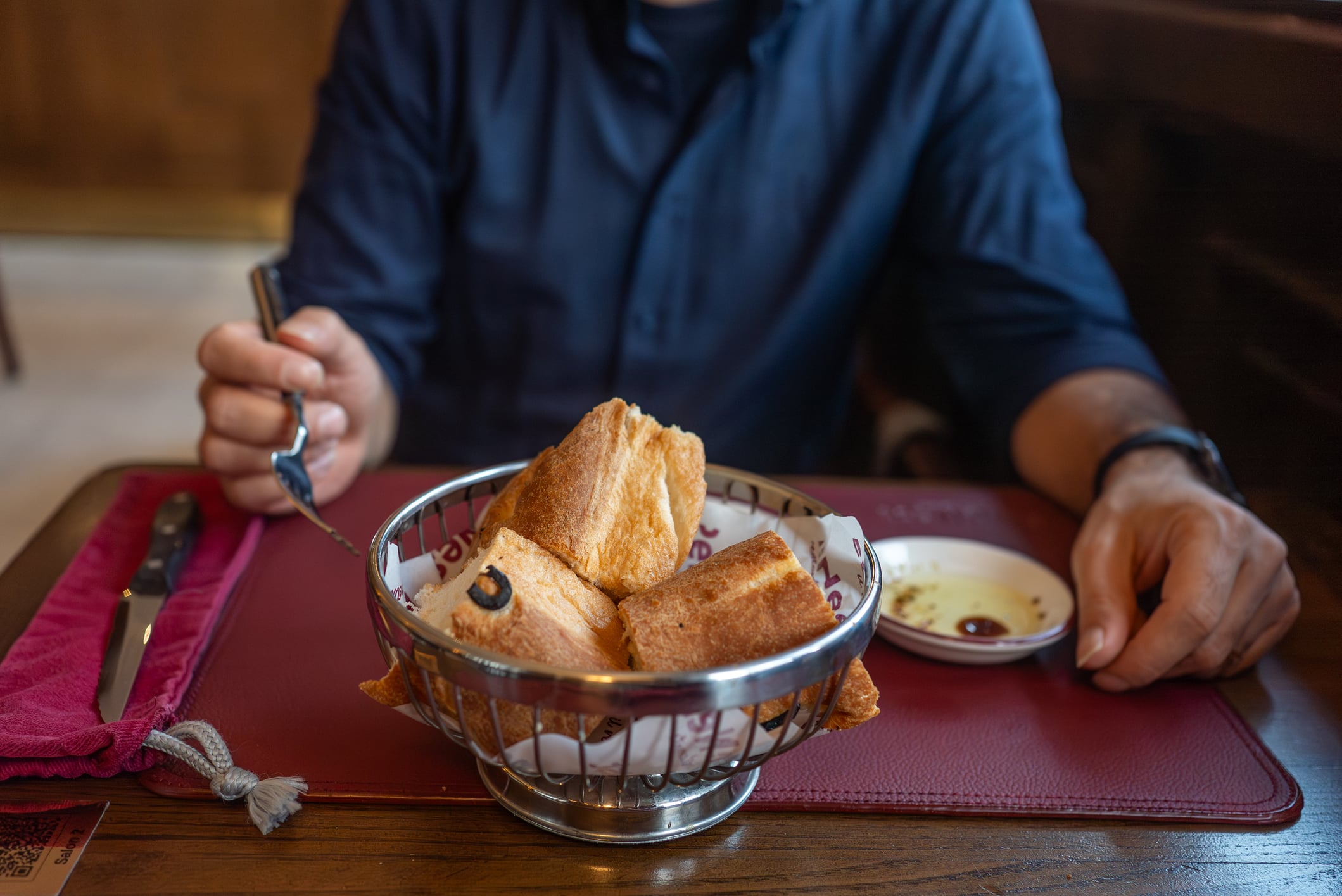 A person sitting at a table with a metal basket of bread rolls and a small dish of oil and vinegar.