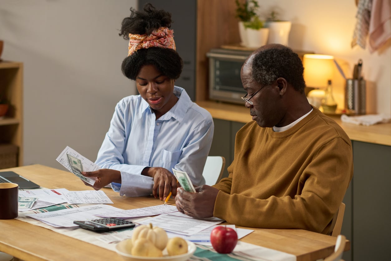A young woman and an older man sit at a table reviewing financial documents and counting cash.