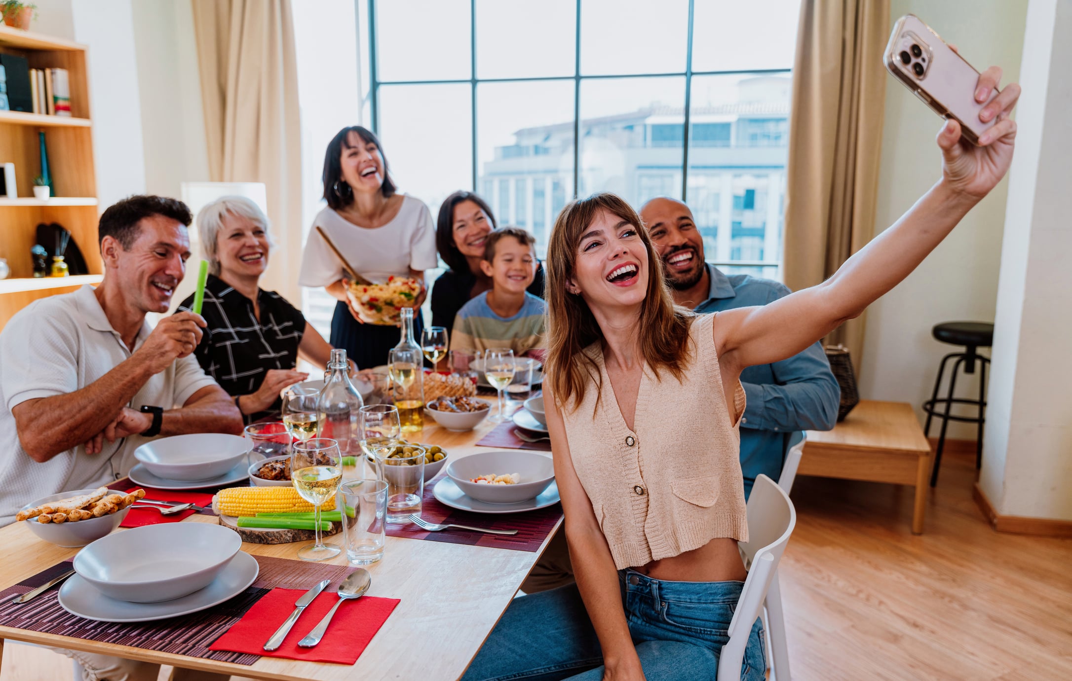 A group of friends and family laughing together at a dining table while a woman in the foreground takes a selfie with her phone.
