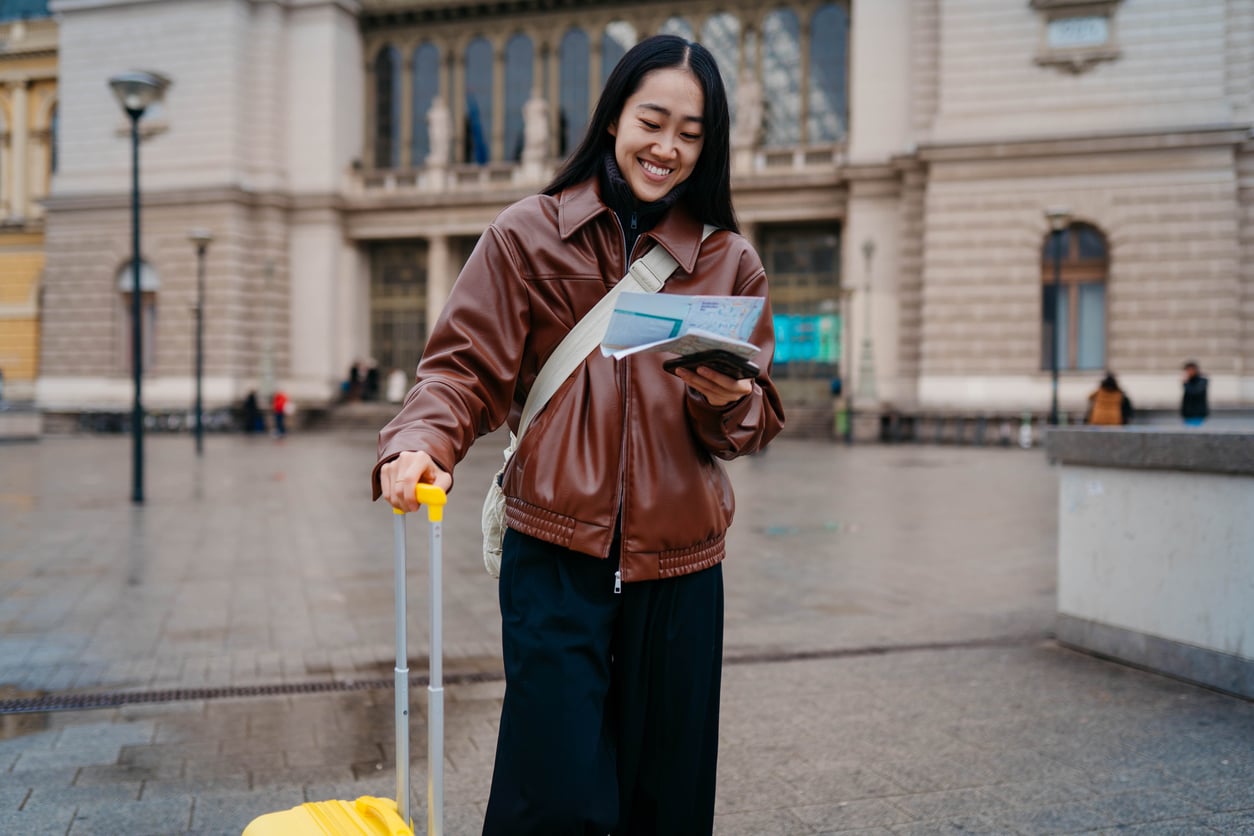 A smiling woman in a brown leather jacket holding a travel document and passport while standing next to a yellow rolling suitcase in front of a historic building.