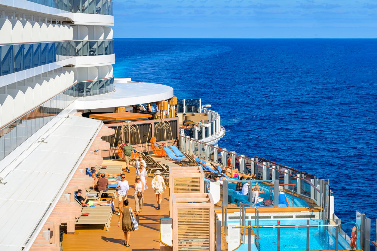 People relaxing on the tiered sun deck of a cruise ship alongside a pool overlooking the ocean.