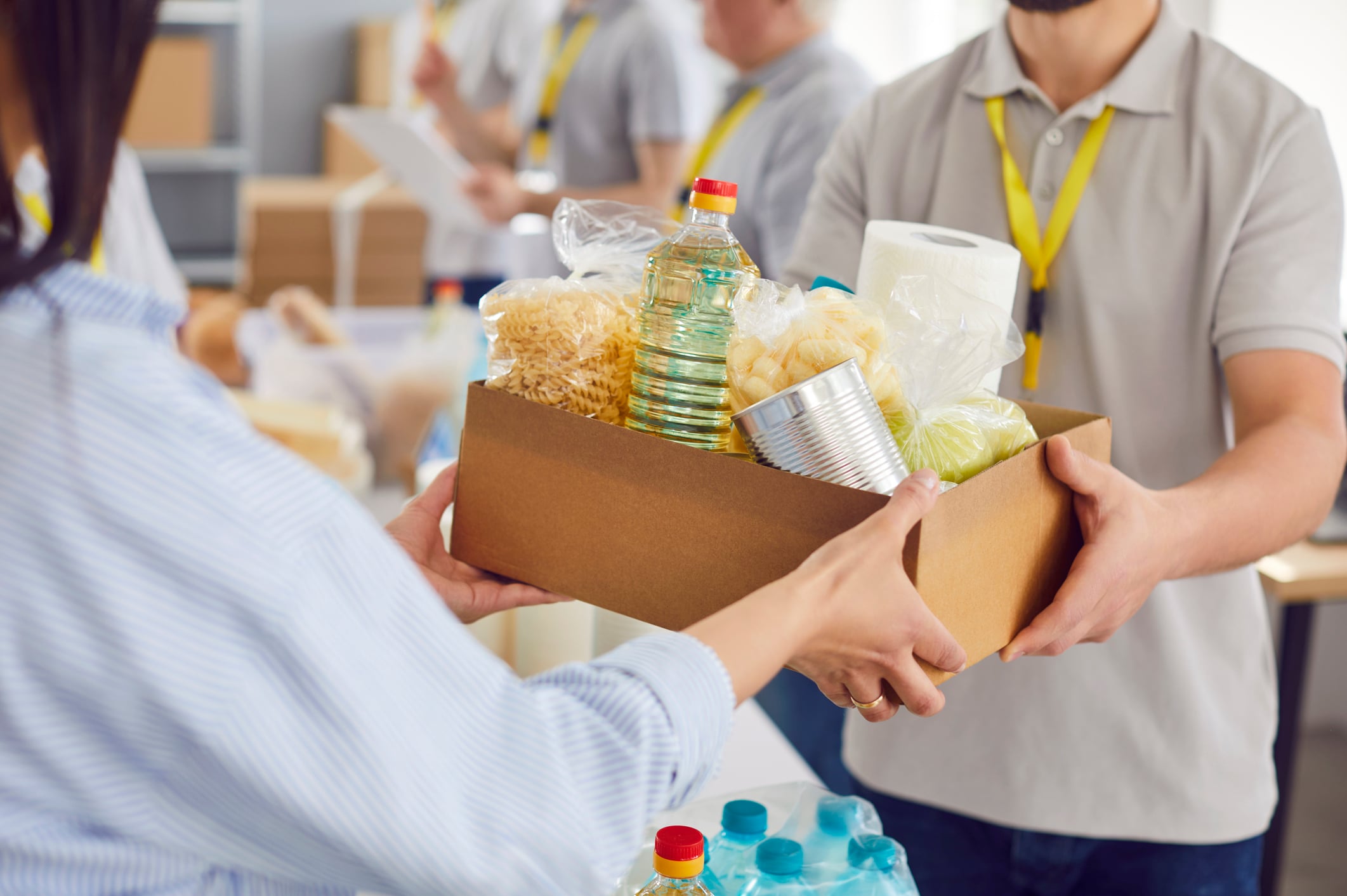A volunteer hands a cardboard box filled with pantry staples, including pasta, cooking oil, and canned goods, to a recipient.