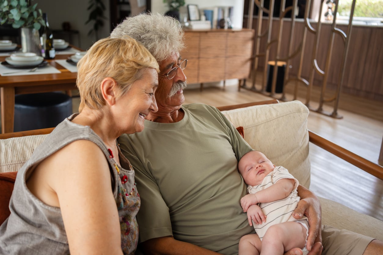 An older couple sits on a sofa, looking down affectionately at a sleeping baby resting in the man’s arms.
