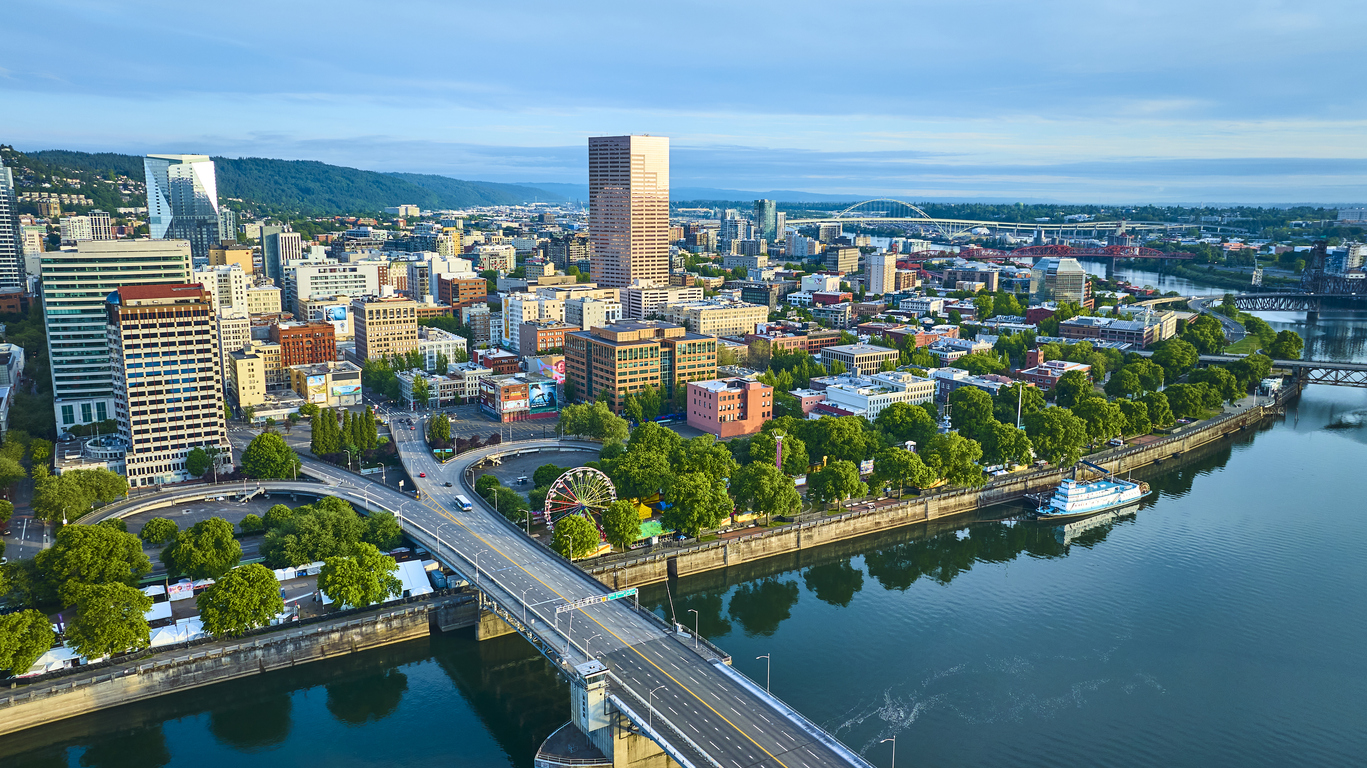 Aerial view of downtown Portland, Oregon, featuring city buildings, a bridge over the Willamette River, lush trees, and a riverboat docked along the waterfront under a clear blue sky.