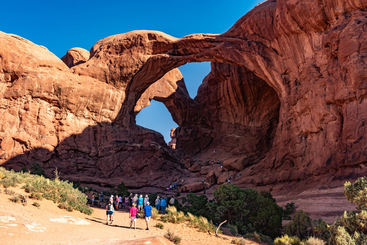 A group of people standing on a sandy path looking up at the large sandstone arches of Double Arch in Arches National Park.