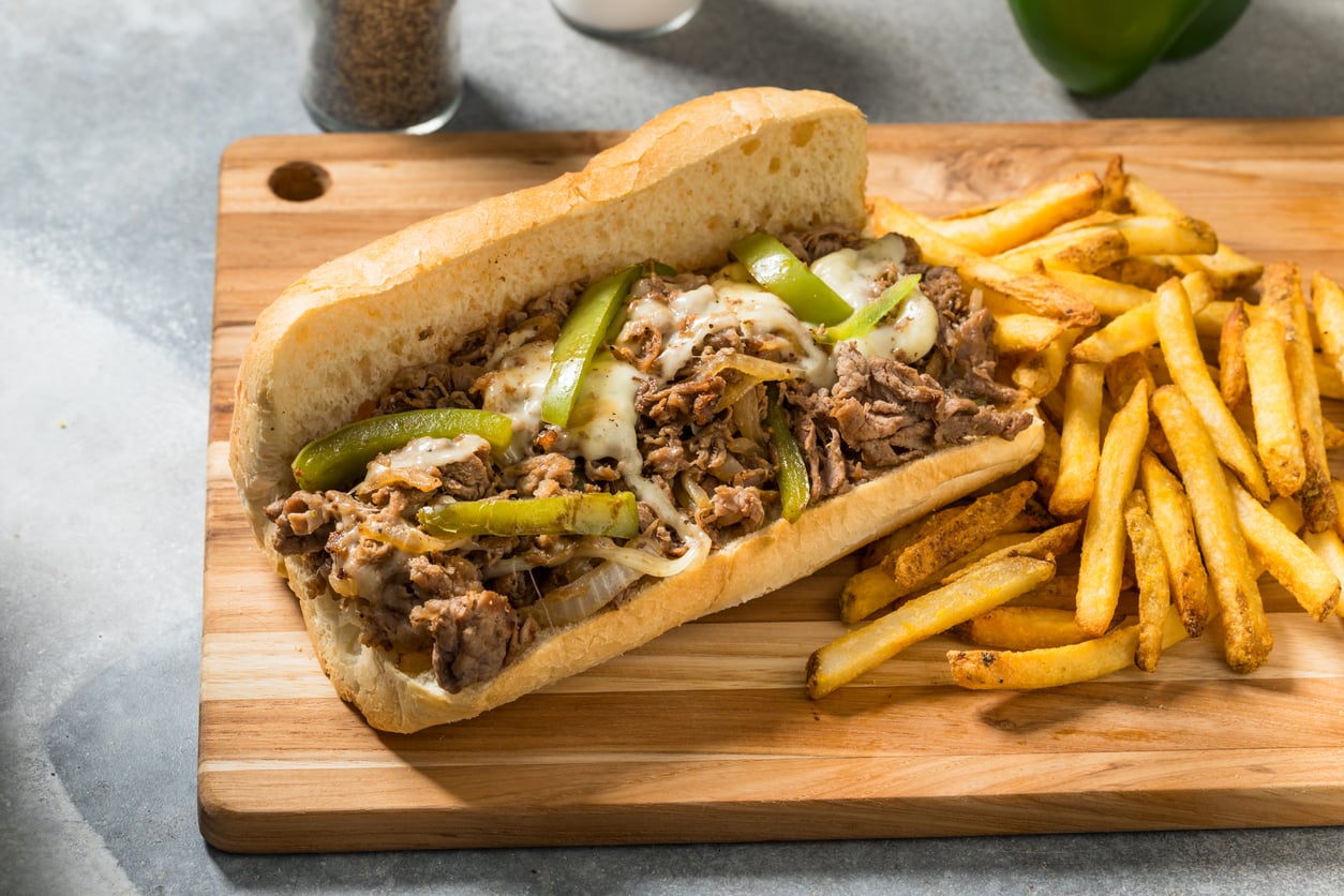 Philly cheesesteak sandwich with green peppers on a wooden board next to a serving of french fries.