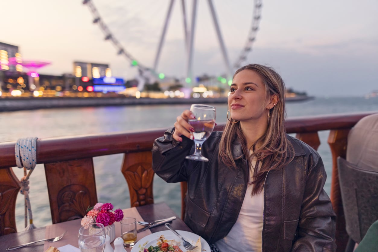 A woman in a dark leather jacket raises a glass of water while dining at an outdoor table with a large Ferris wheel in the background.