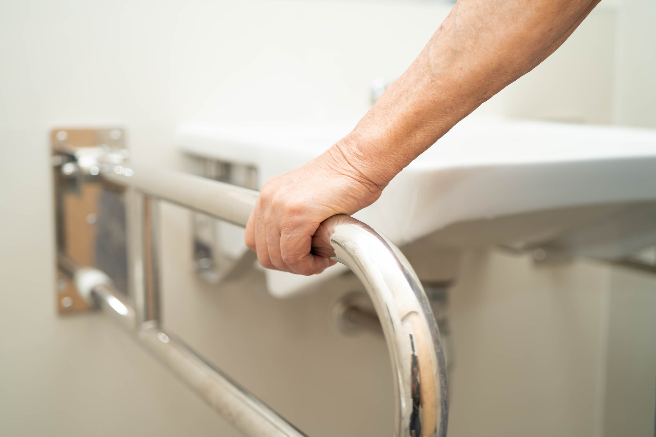 An older person’s hand grips a stainless steel grab bar mounted to the wall next to a bathroom sink, an essential home improvement for accessibility.