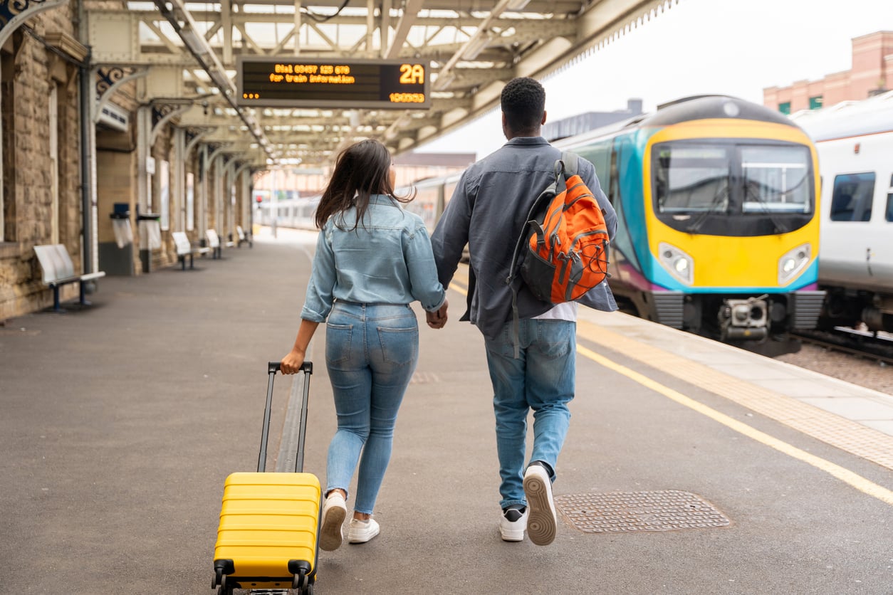 A couple holding hands walks along a train platform toward a waiting train, with the woman pulling a yellow suitcase and the man wearing an orange backpack.
