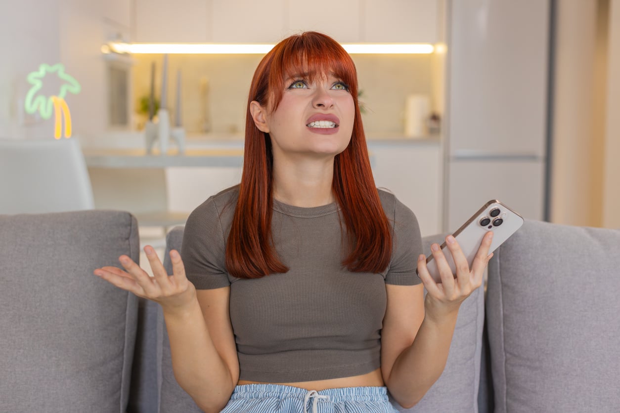 A woman with red hair sitting on a couch looks upward with a frustrated expression while holding a smartphone.