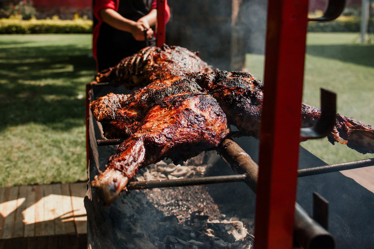 Large cuts of meat roasting over hot coals on a metal spit.