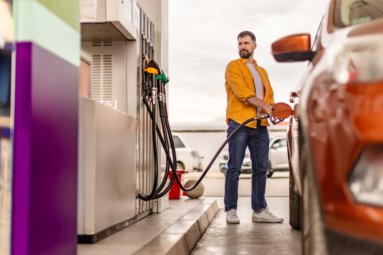 A man in a yellow shirt pumping gas into his orange car at a fuel station.