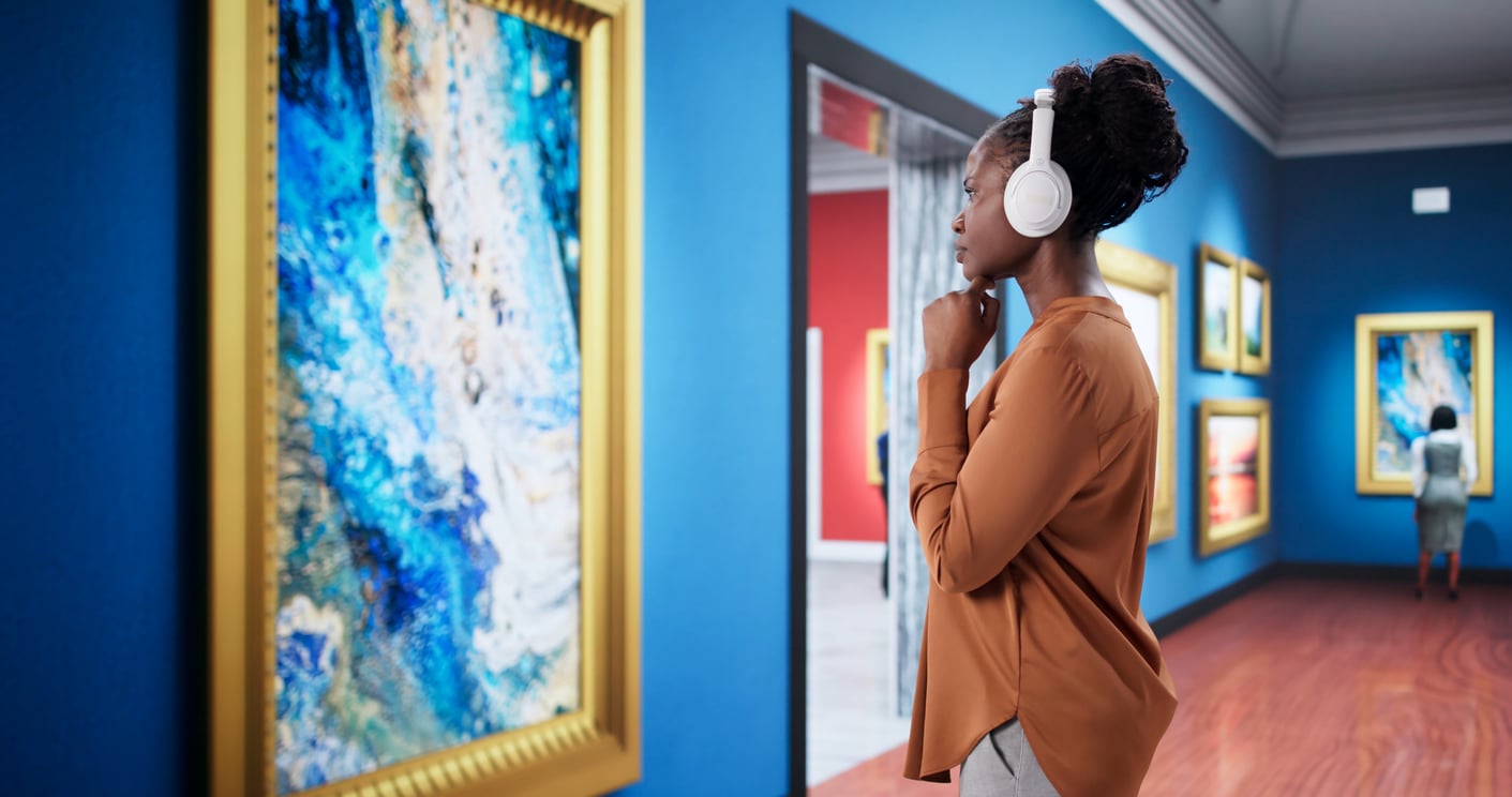 A woman wearing white over-ear headphones contemplates a framed abstract painting in an art gallery.