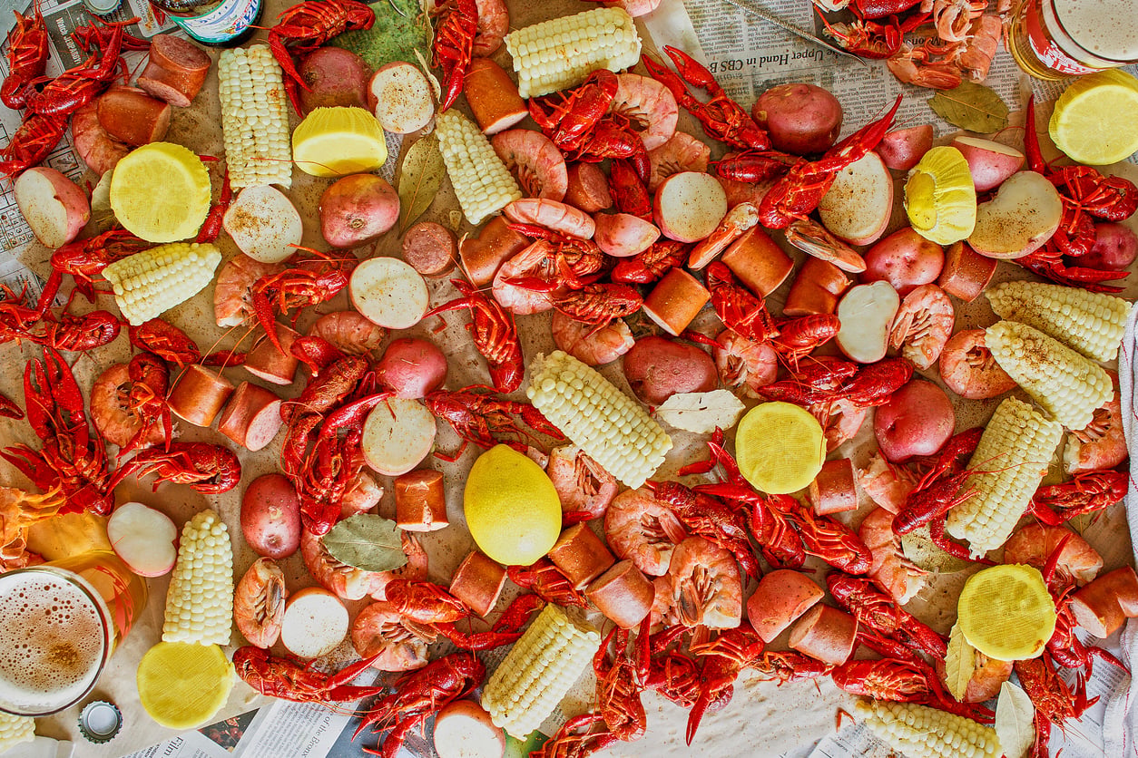 A seafood boil featuring crawfish, shrimp, sliced sausage, corn on the cob, and red potatoes spread across a newspaper-covered surface, with lemon halves and a glass of beer visible.