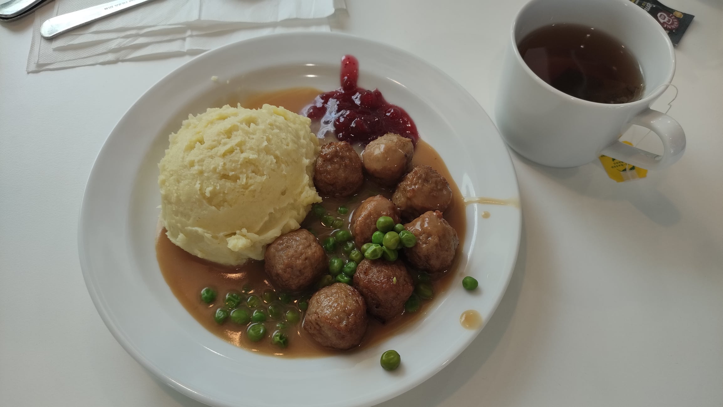 A plate of Swedish meatballs with mashed potatoes, gravy, green peas, and lingonberry jam, next to a mug of tea.