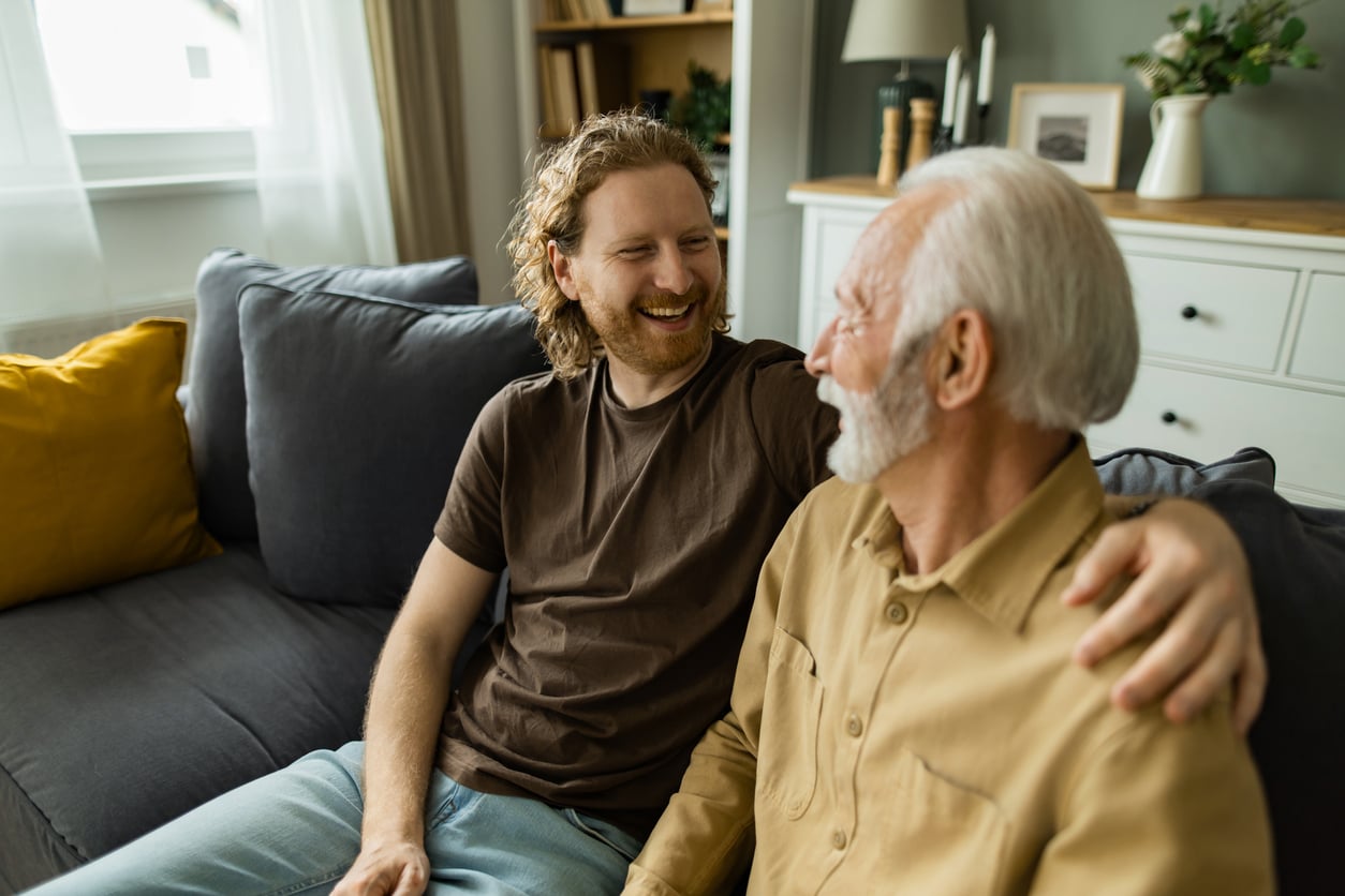 A smiling young man with curly hair has his arm around an older man with a white beard as they sit together on a sofa.
