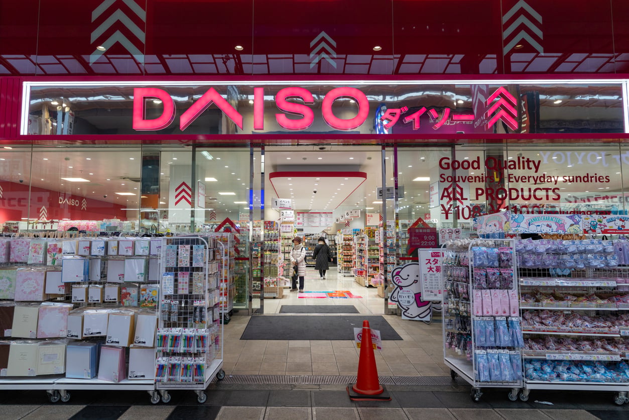 The storefront of a Daiso variety store, with racks of merchandise displayed outside the entrance and a large pink sign above the door.