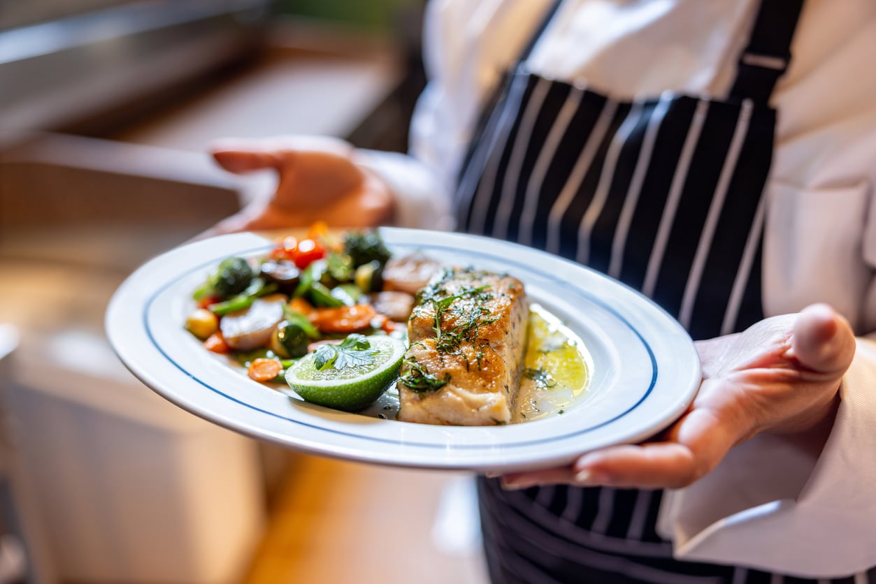 A chef in a striped apron holds a plate featuring a piece of cooked white fish, a lime wedge, and sautéed vegetables, perfect for restaurant menu tips.