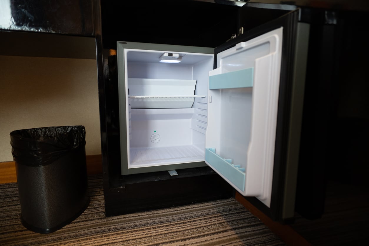 An open, empty mini-fridge built into a cabinet next to a black trash can.