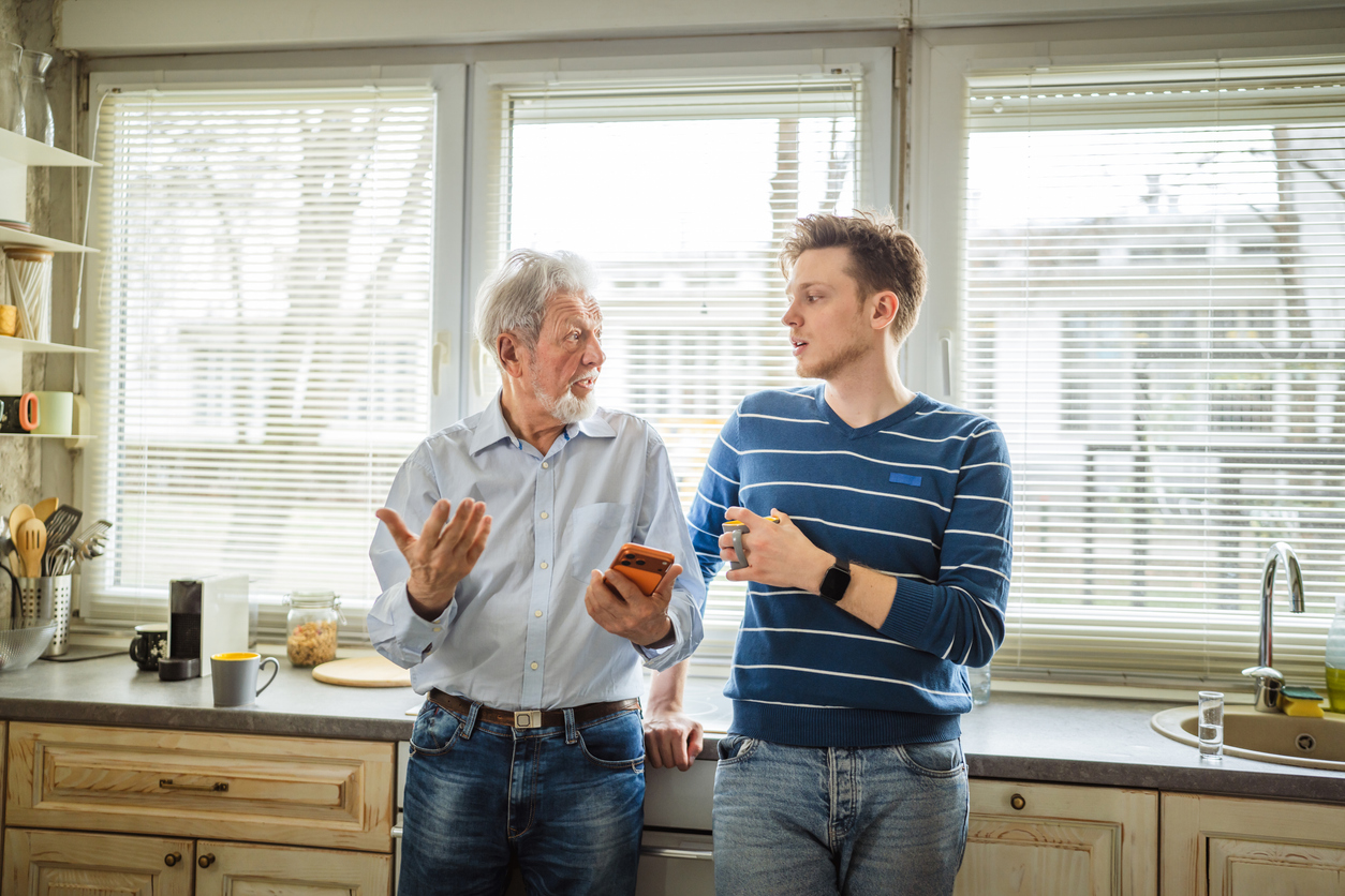 An older man holding a smartphone and a younger man holding a mug talk while standing in a kitchen.