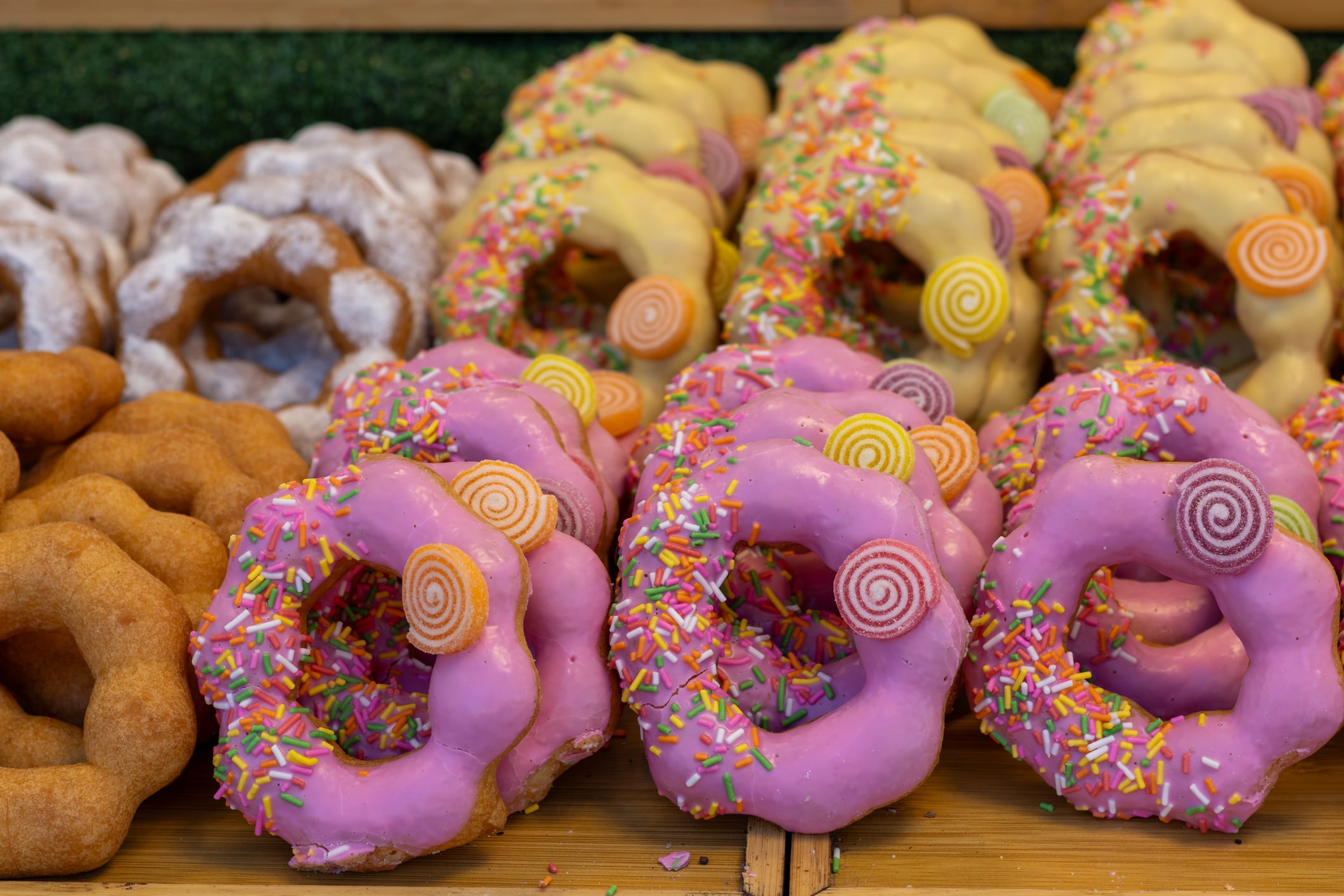 Rows of assorted ring-shaped donuts, including pink-frosted with sprinkles and candy, white-frosted, and plain sugar-dusted varieties.