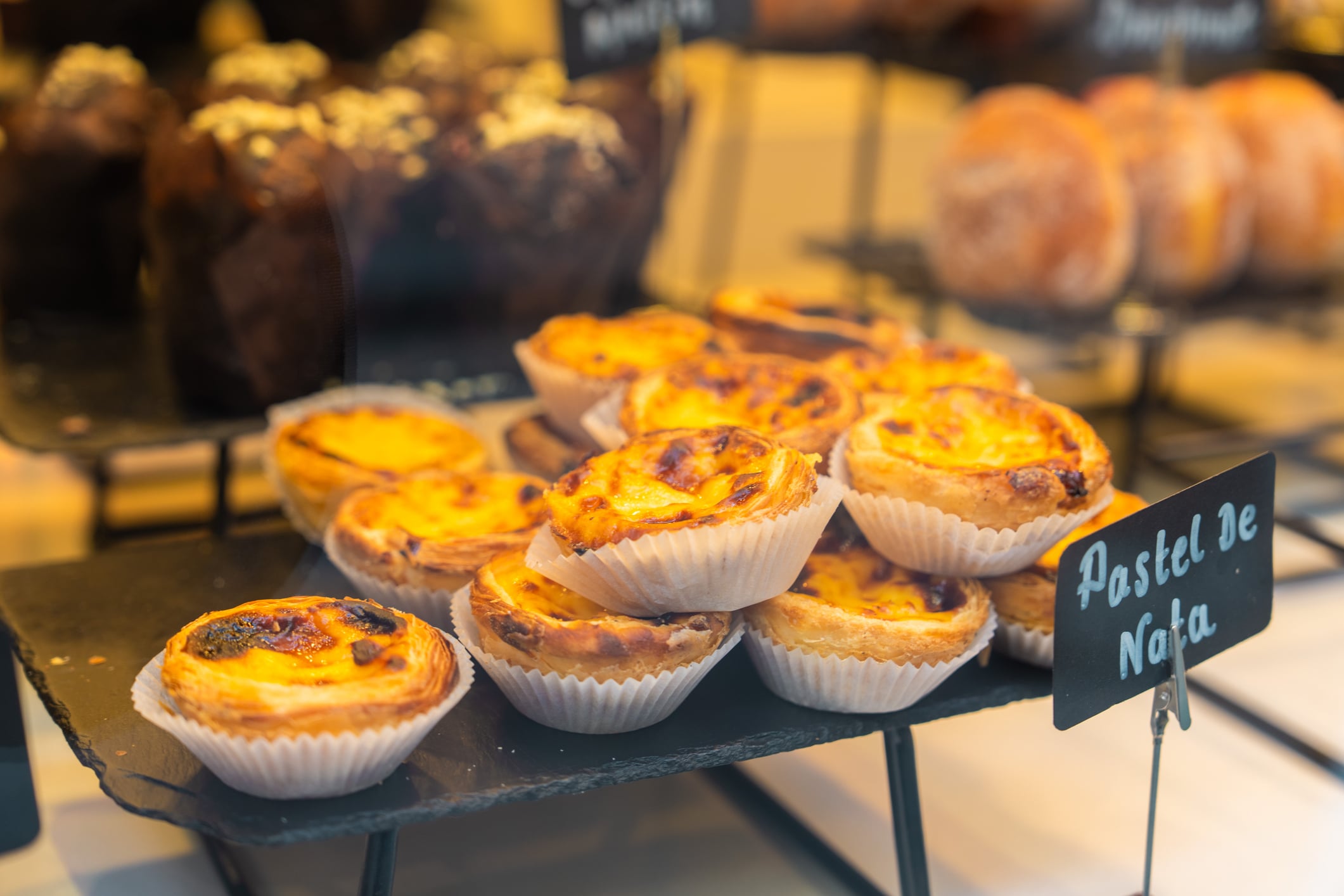 A display of several egg custard tarts in paper liners on a black slate tray, with a sign reading “Pastel De Nata.”