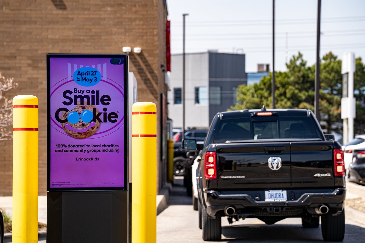 A digital menu board at a drive-thru displays an advertisement for “Smile Cookie” charity cookies, featuring the dates April 27 to May 3 and the name ErinoakKids, with a black pickup truck parked in the lane in front of it.