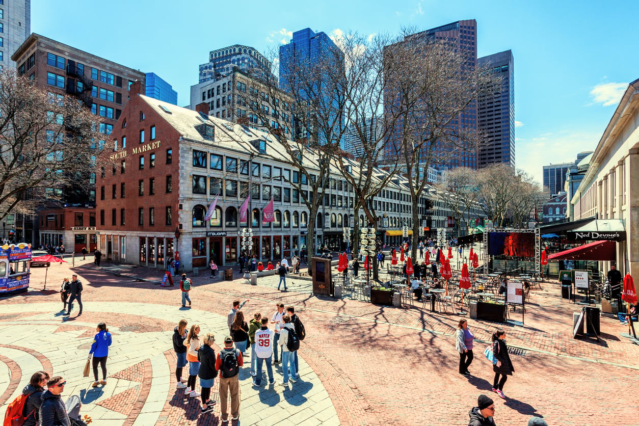 A brick-paved plaza at Faneuil Hall in Boston with people walking, scattered outdoor tables, and surrounding historic brick buildings and modern skyscrapers.
