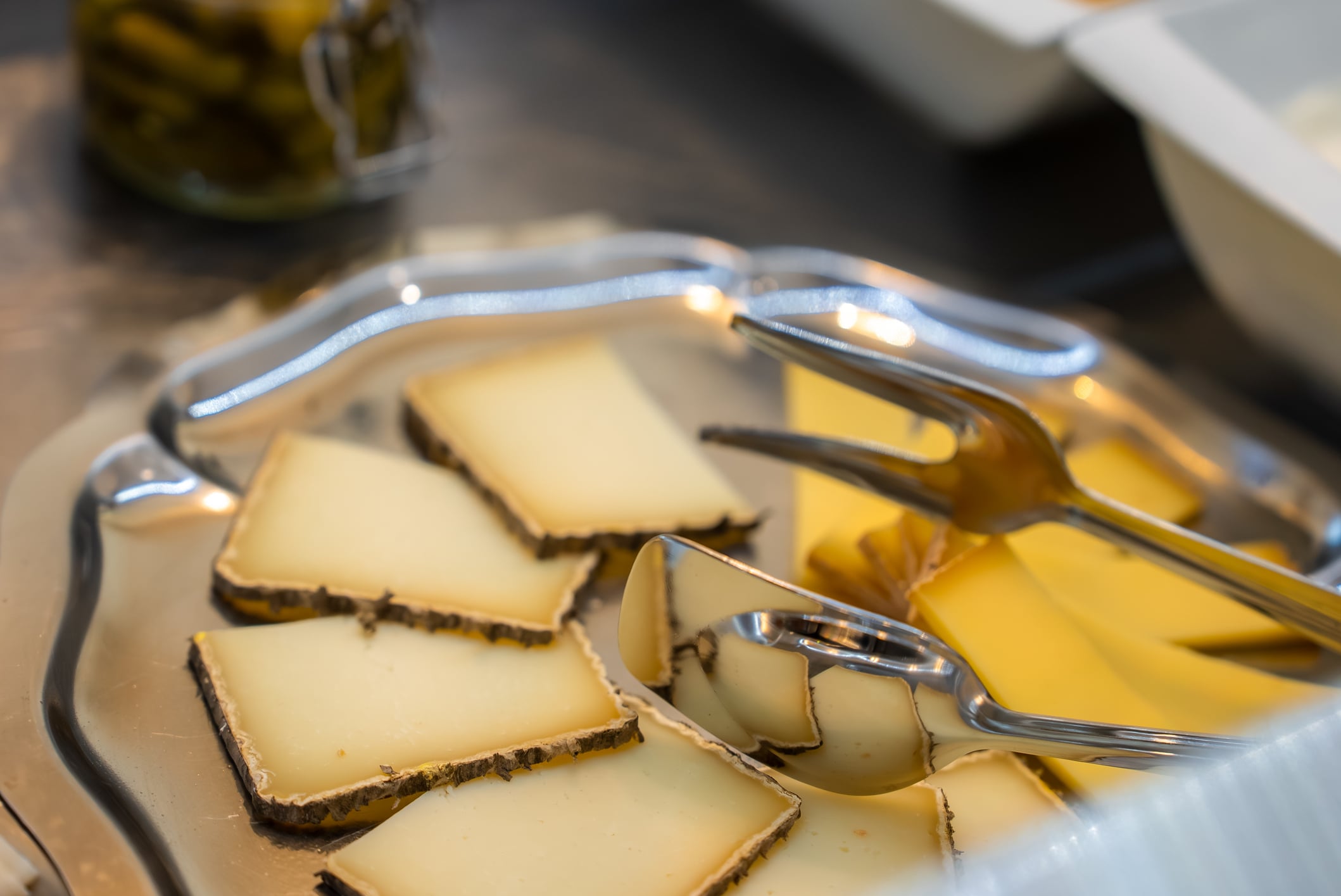 Slices of cheese with dark rinds on a silver platter with a serving fork and tongs.